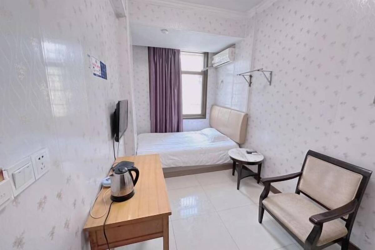 Una habitación pequeña y luminosa con una cama, una silla, una mesa con un hervidor y un televisor.
