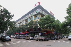 A multi-story hotel with a prominent red sign on the roof, a parking lot with several cars, and a street view with trees and storefronts.
