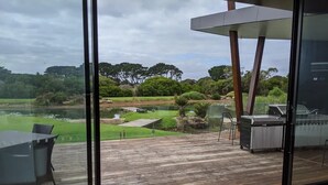 Vue depuis une pièce dotée de grandes fenêtres donnant sur une terrasse aménagée avec des meubles d’extérieur, un barbecue et un étang.