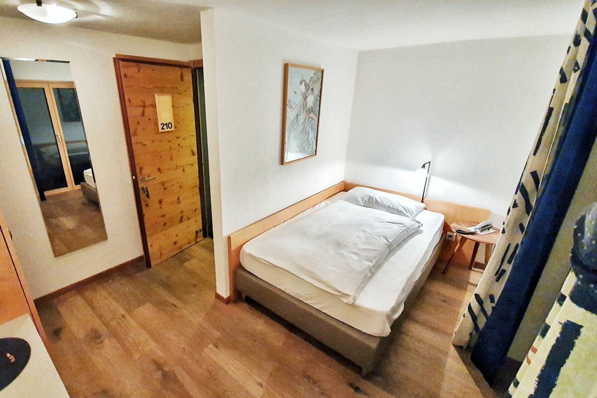 A hotel room with a wooden floor, a bed with white bedding, a bedside table with a lamp, a door with a number, and a mirror on the wall.