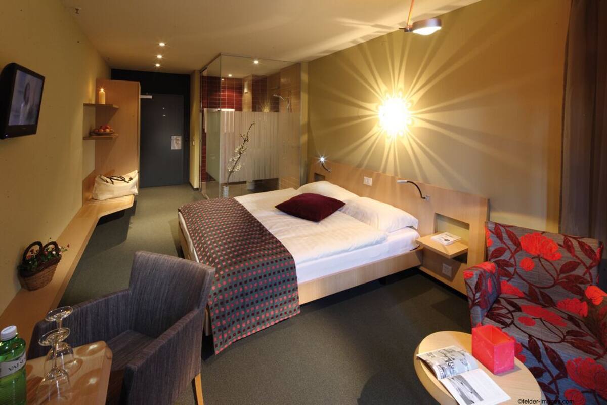 A hotel room with a bed, a chair, a table with a bottle, a lamp, and a TV on the wall.