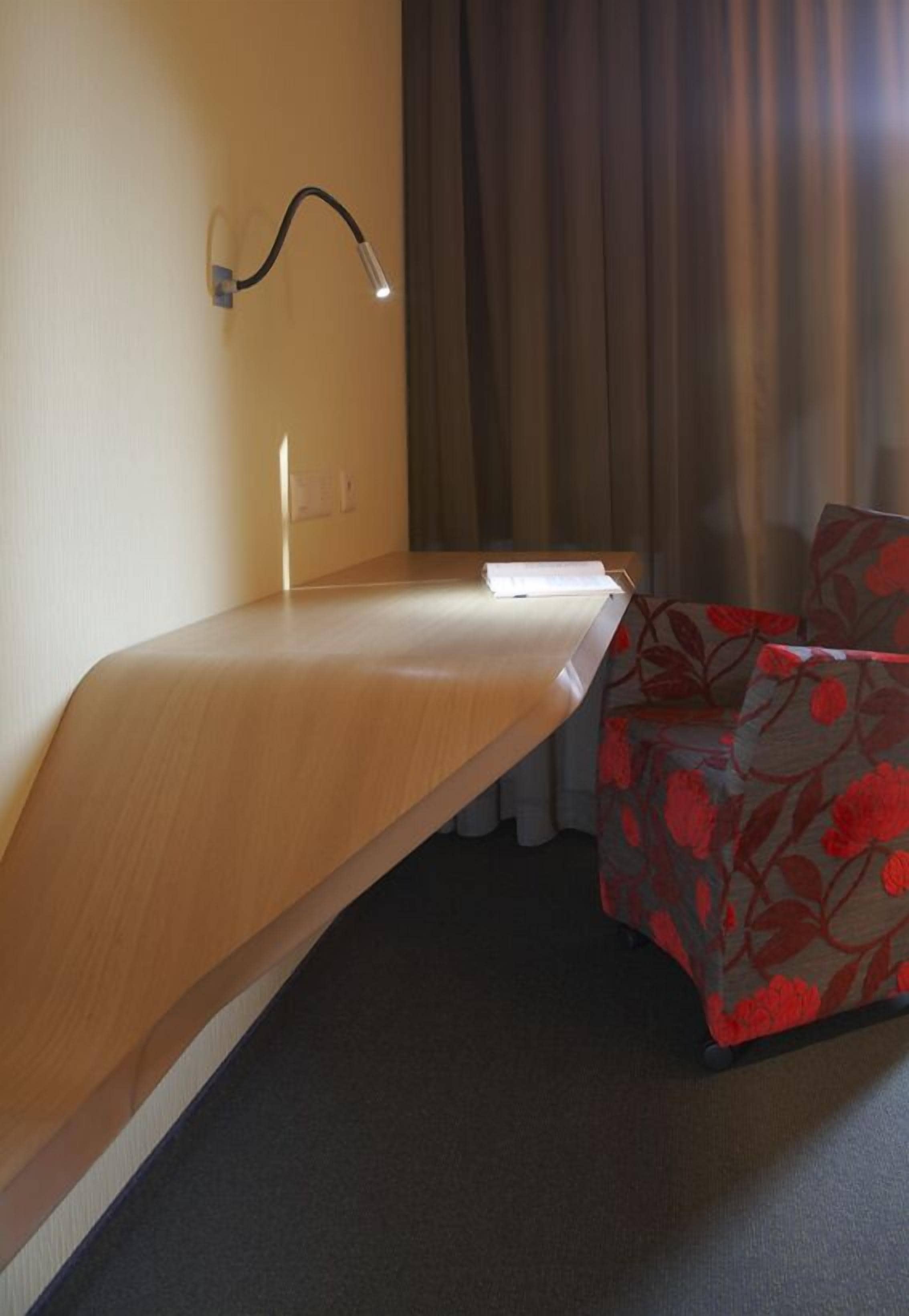 A hotel room with a wooden desk, a chair with a floral pattern, a wall-mounted lamp, and a window with curtains.
