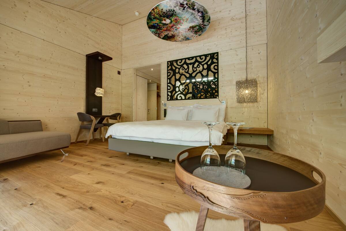 A modern room with a wooden floor, a bed with white linens, a round wooden coffee table with two wine glasses, a gray sofa, and a decorative wall piece.