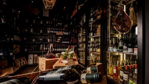 Un bar tenuemente iluminado con estantes llenos de botellas diversas, una barra de madera con vasos, y un mostrador con exhibición de carnes colgantes.