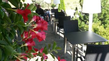 An outdoor patio area with tables and chairs, surrounded by greenery and a covered section.
