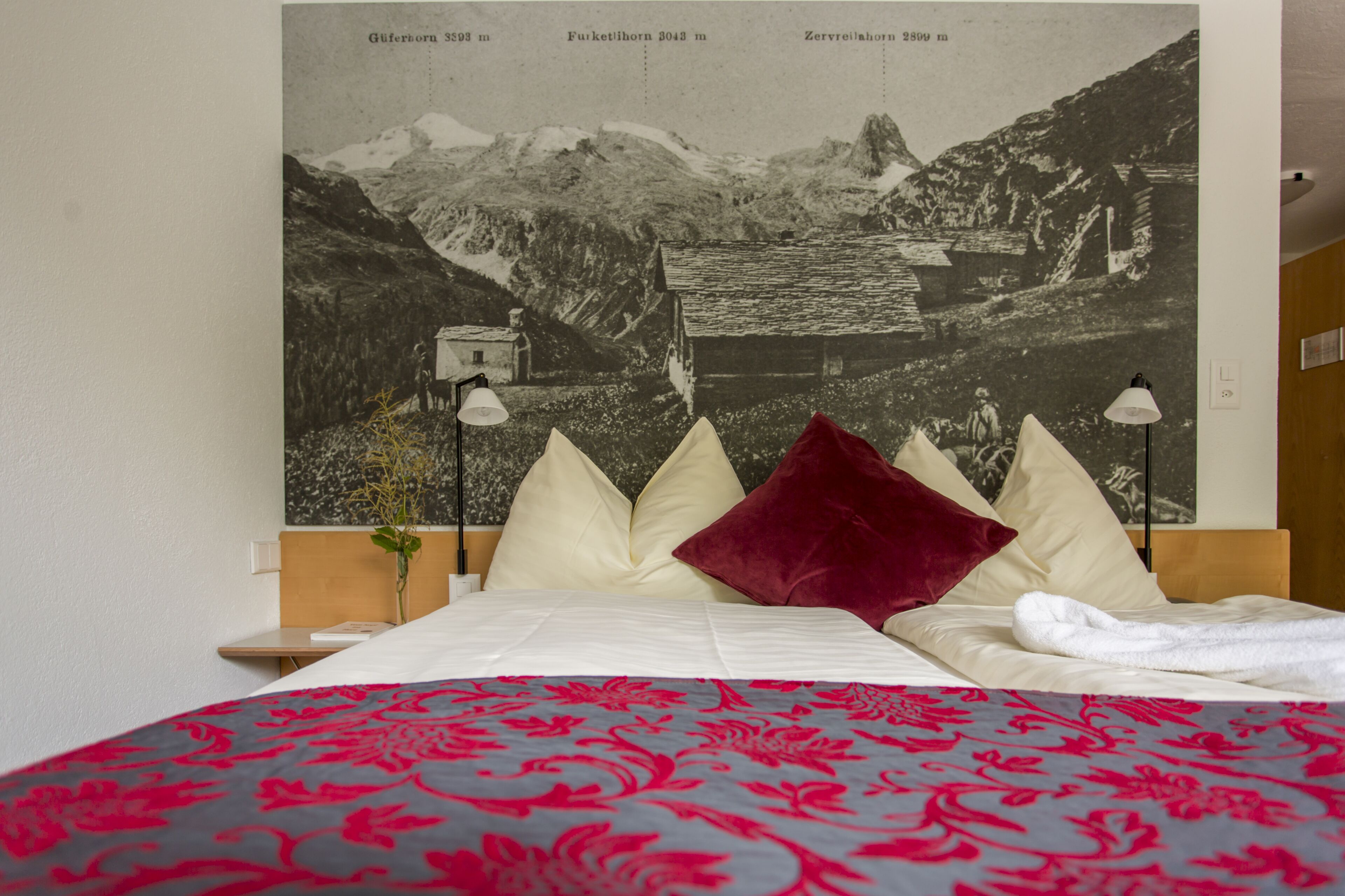 A bed with a patterned bedspread, a red pillow, and a framed landscape photograph above it.