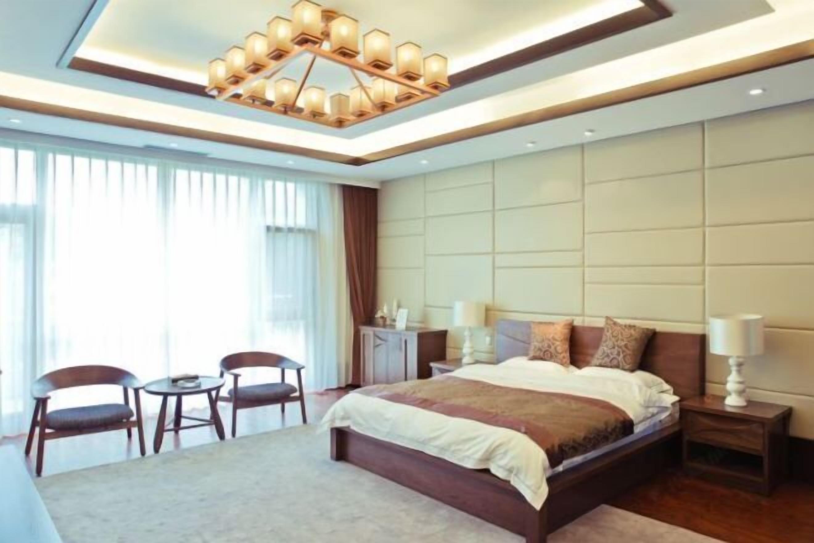 A modern hotel room with a large bed, bedside tables, a chandelier, and a view of the city.