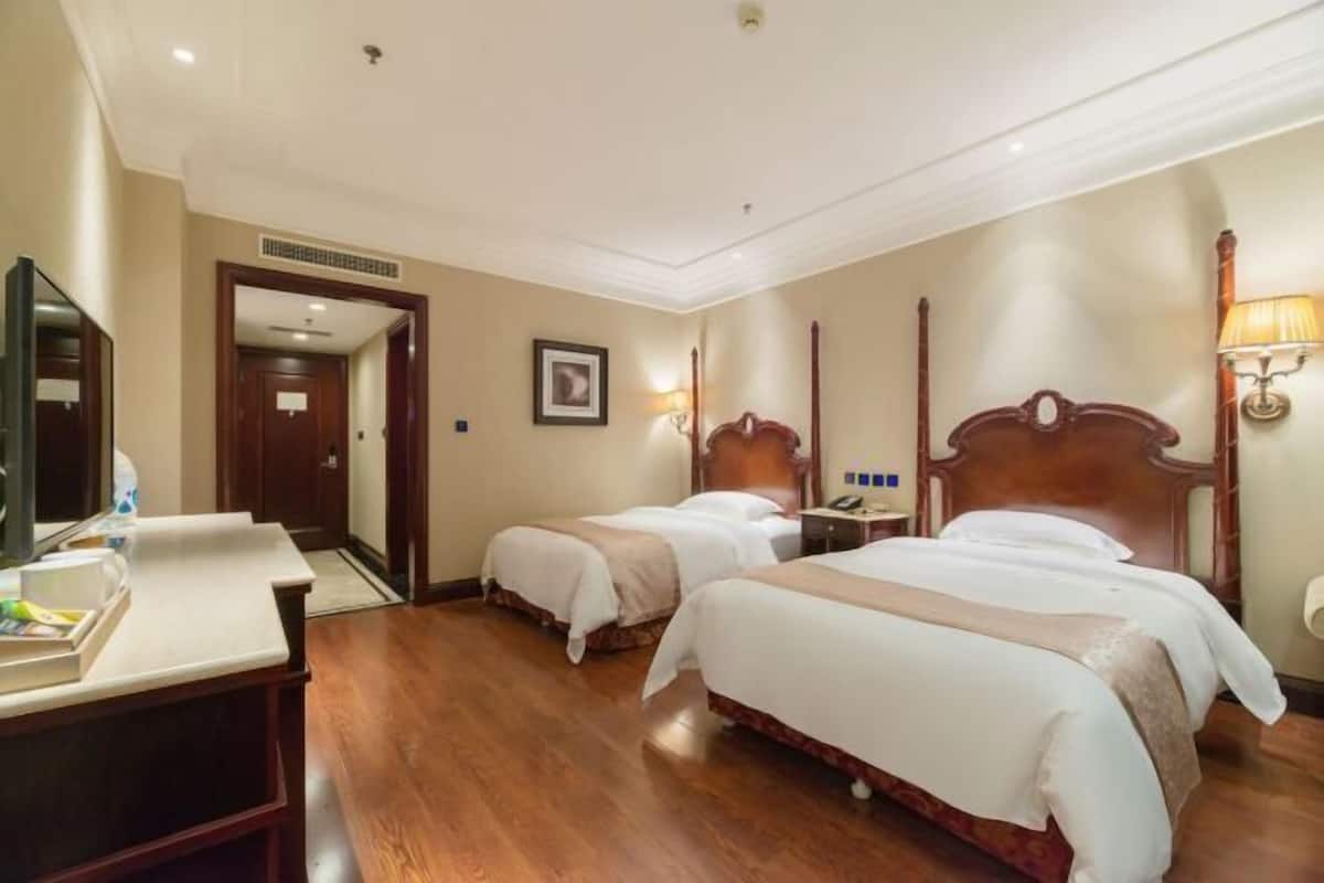 A hotel room with two beds, a desk, a mirror, a door, and a wall-mounted lamp.