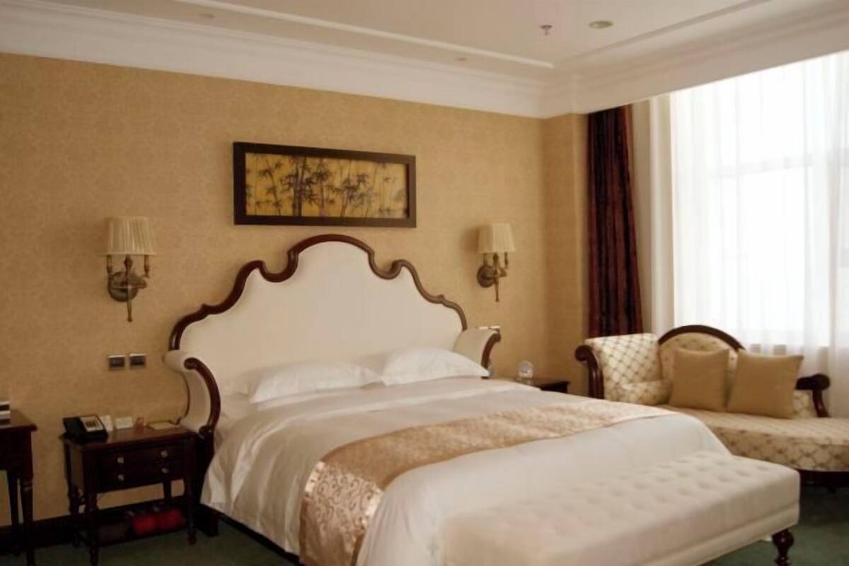 A hotel room with a large bed, a bedside table, a chair, a wall-mounted lamp, and a window with curtains.