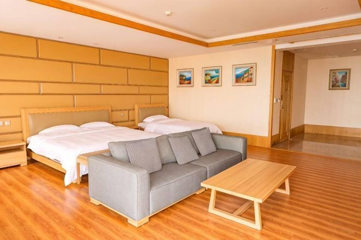 A hotel room with two beds, a grey sofa, a wooden table, and artwork on the walls.