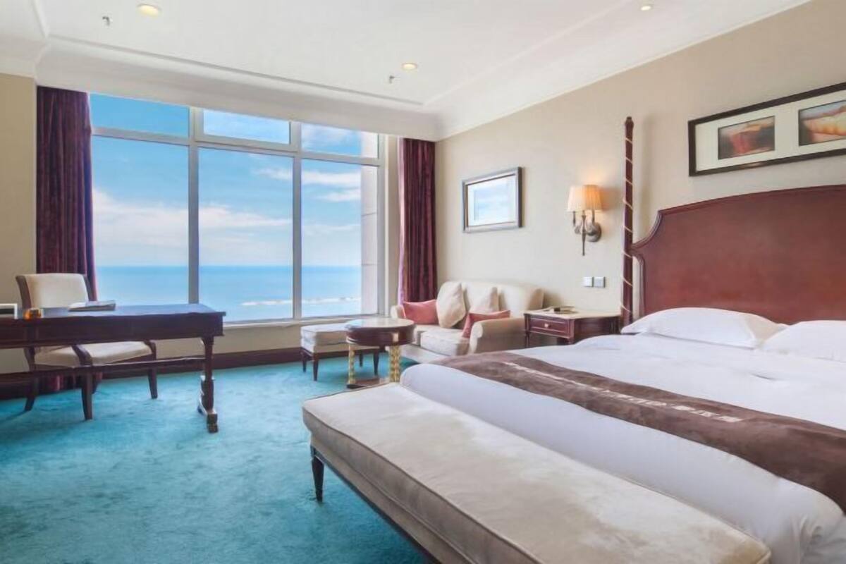 A hotel room with a large window, a bed, a desk, a chair, and a small table.