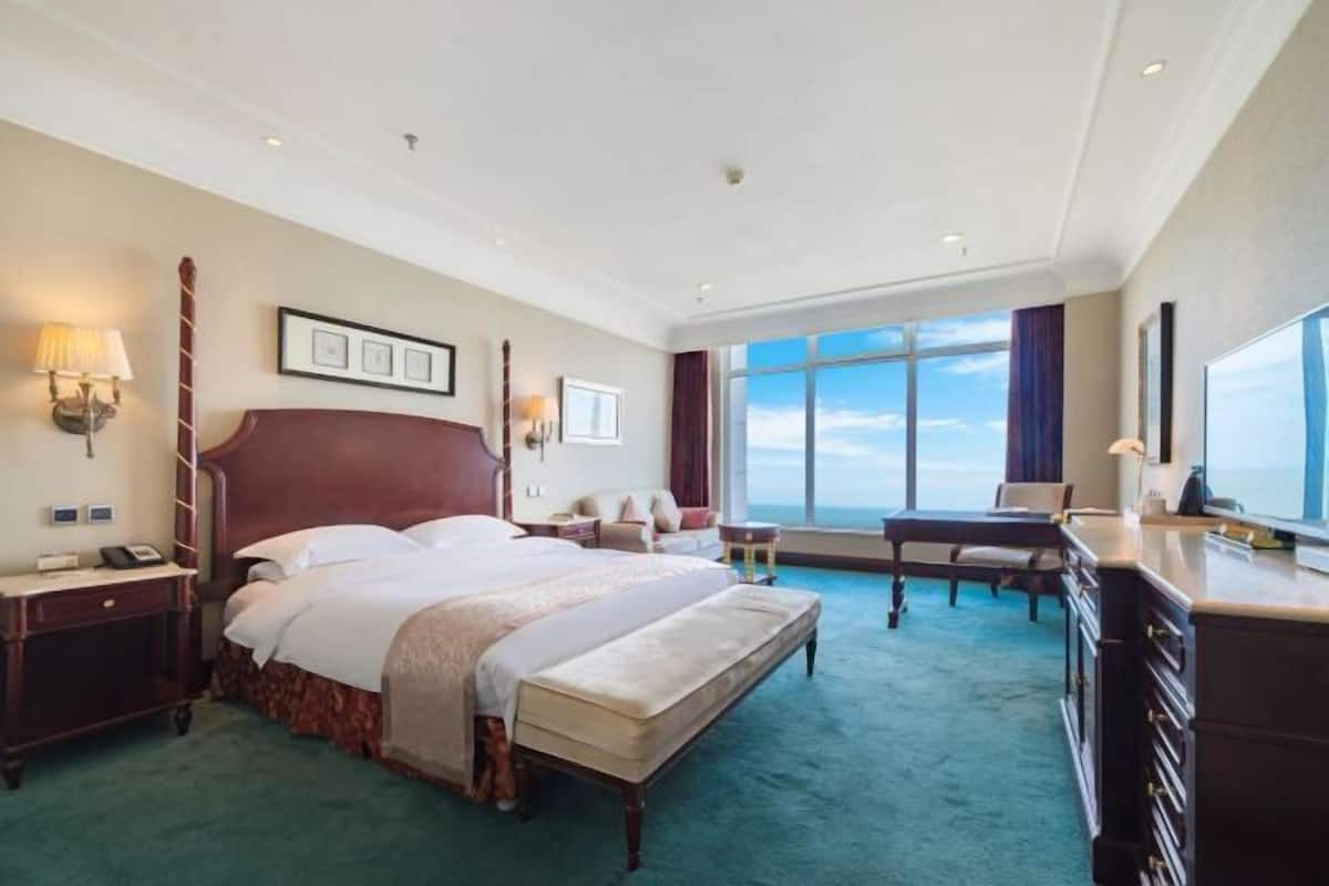 A hotel room with a large bed, a desk, a chair, a nightstand, a lamp, and a large window offering a view.