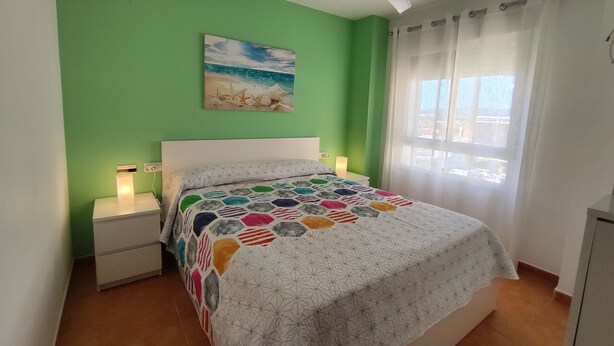 Een slaapkamer met een groene muur, een schilderij met een strandthema, een plafondventilator en een bed met een kleurrijke quilt.