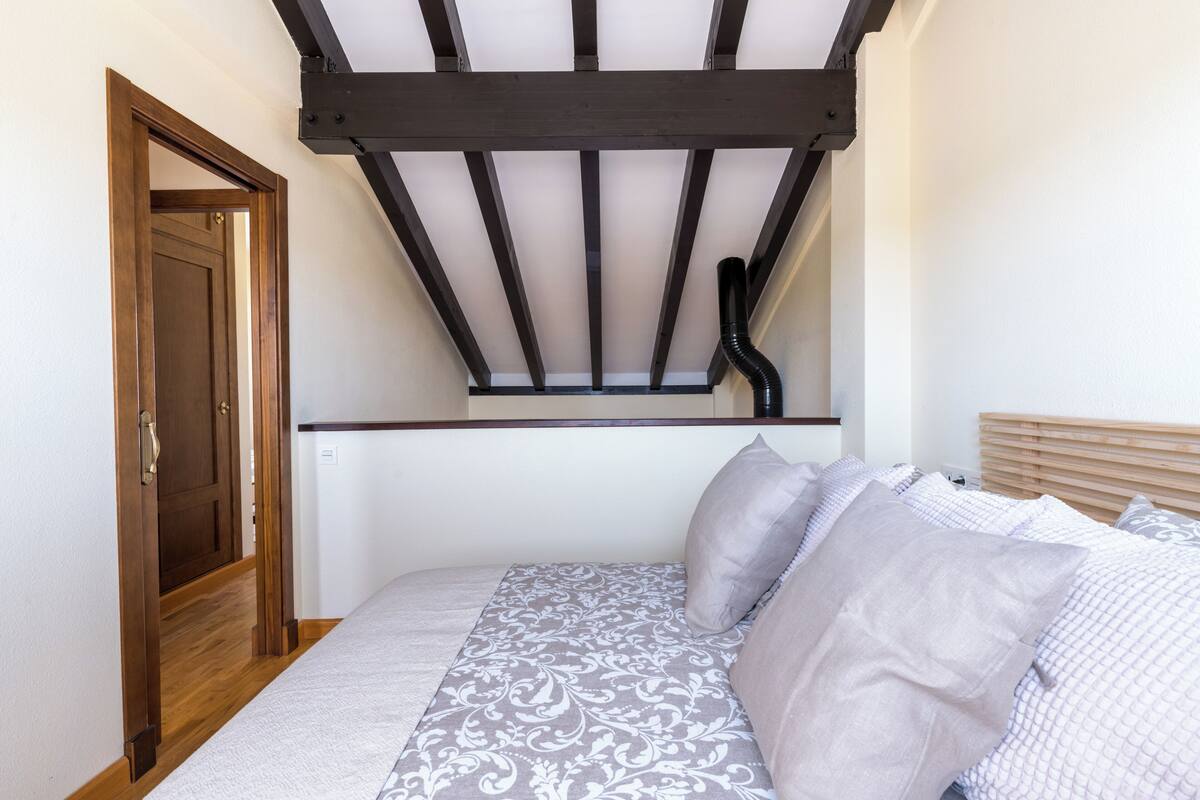 Ein Schlafzimmer mit schrägen Wänden, einer Holztür, einem Bett mit gemusterten Betttextilien und einem Kopfteil.