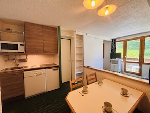 Eine kleine Küche mit Mikrowelle, Spüle und Schränken, ein Esstisch mit Stühlen und ein Fenster mit Blick auf Grünflächen.