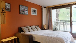 A bedroom with a bed, a wooden nightstand, a window with curtains, and a view of outdoor furniture.