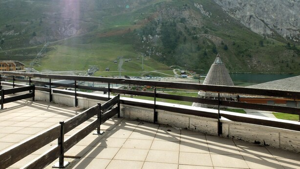 A mountain view from a balcony with a wooden railing, a ski lift in the background, and a lake.