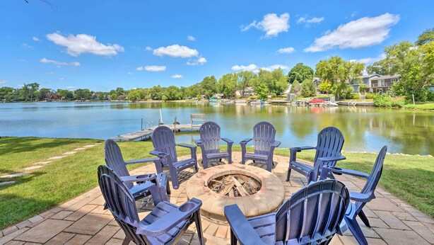 A fire pit surrounded by Adirondack chairs on a patio with a view of a lake and houses in the background. - Lakefront Oasis w/ Boat Dock, Fire Pit, Grill (Bristol)