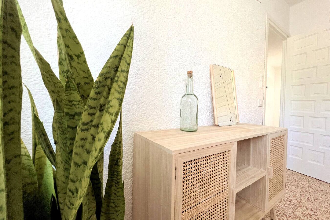 Une console en bois, sur laquelle sont posés un flacon et un miroir, est adossée à un mur blanc, à côté d’une grande plante.