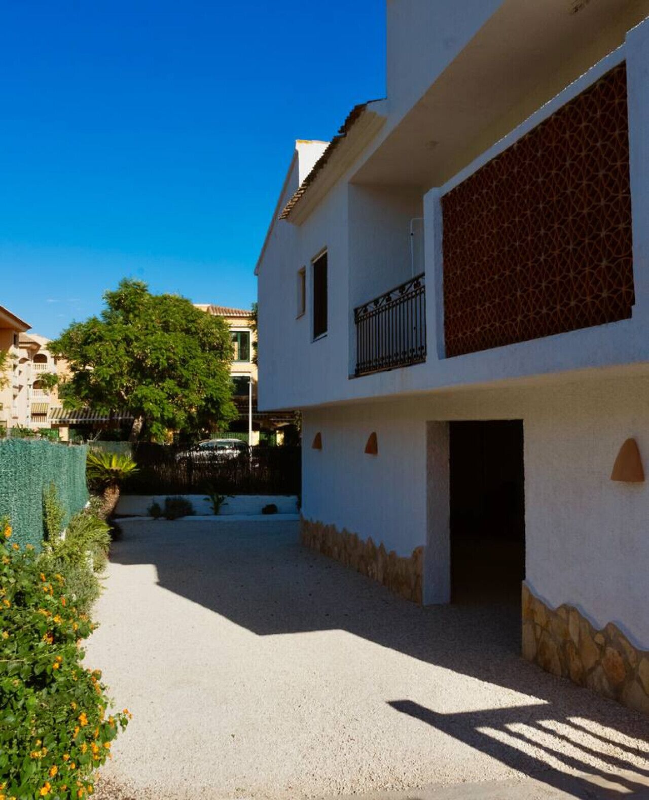 A residential building with a paved courtyard, a visible entrance, and a tree in the background.