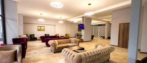 A spacious living area with multiple seating options, including a large beige sofa, a smaller armchair, and a red armchair. There is a television mounted on the wall, and the room features a patterned wall and a wooden door in the background.