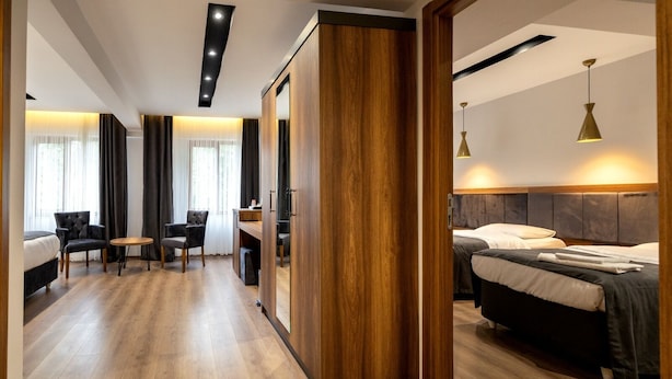 A modern hotel room with a wooden floor, a desk with chairs, a bed with a headboard, and a large mirror. - Turkler Hotel (Trabzon)