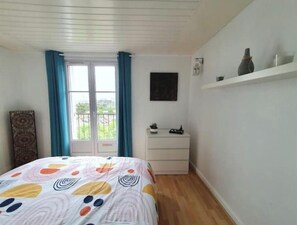 A bedroom with a bed, a dresser, a window with curtains, a small shelf with decorative items, and a wall-mounted light. - L'Escapade Océane - downtown Le Havre (Le Havre)