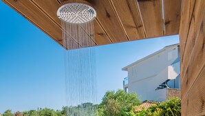 A modern outdoor shower with a wooden ceiling, a white wall, and a view of a house and greenery in the background.