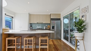 A modern kitchen with a large island, wooden stools, a refrigerator, and a view of the outdoors.