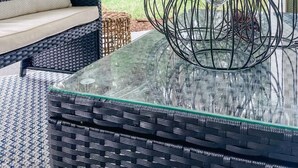 A glass-top table with black metal candle holders, wicker furniture, and a view of trees and a house in the background.