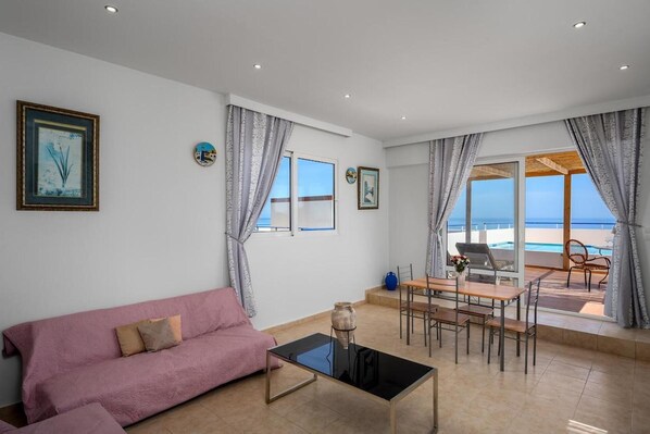 A living room with a pink sofa, a glass coffee table, and a dining area with a wooden table and chairs. There are framed pictures on the wall and a view of the beach through large windows. - Exquisite Retreat For Two (Rhodes)