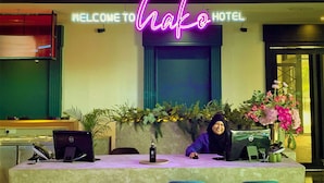 A reception desk with a neon sign reading "Welcome to Hako Hotel" and a sign below stating "Here to check you in... then out." - HAKO HOTEL JBCC (Johor Bahru)