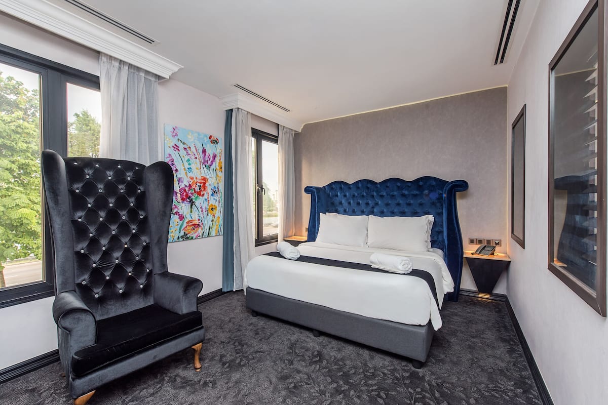 A hotel room with a large bed, a black armchair, a painting, and a view of the outdoors.
