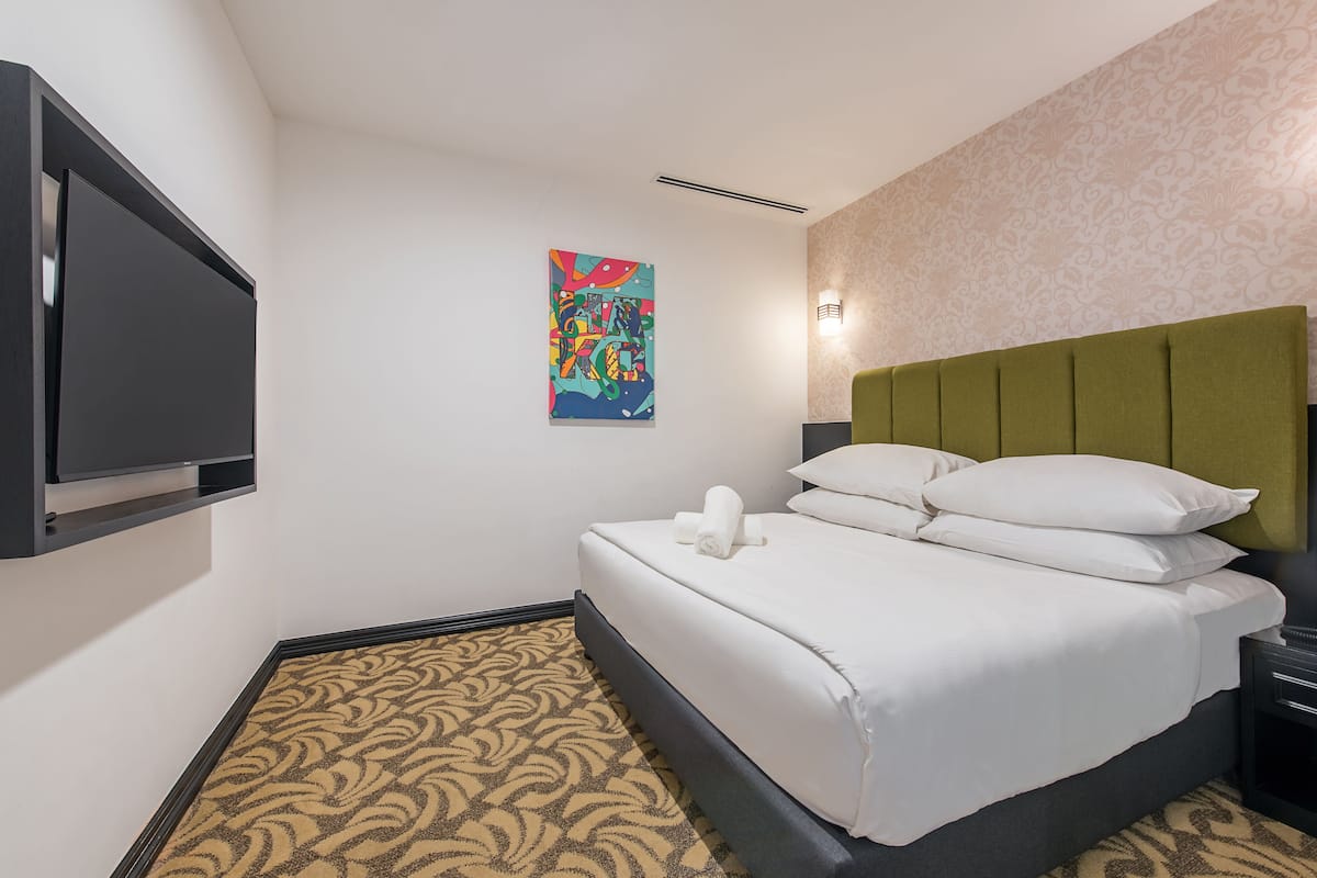 A hotel room with a bed, a flat-screen TV mounted on the wall, a patterned carpet, and a decorative painting.
