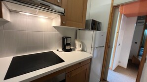 Une cuisine compacte dotée d’une plaque de cuisson à induction noire, d’un réfrigérateur blanc, d’une machine à café et de meubles de rangement en bois.