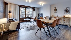 Een moderne woonkamer met een eethoek, grote ramen, een flatscreen televisie en uitzicht op een gebouw.