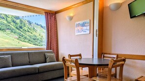 A room with a sofa, wooden chairs, a small table, a window with a view of green hills, and a wall-mounted TV. - Divisible studio at the foot of slopes with balcony, pets allowed (Plagne Aime 2000)
