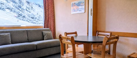 A cozy room with a sofa, wooden dining table, chairs, a TV, and a window with a snowy mountain view.