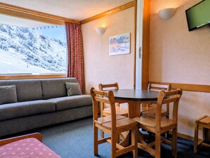 A cozy room with a sofa, wooden dining table, chairs, a TV, and a window with a snowy mountain view.
