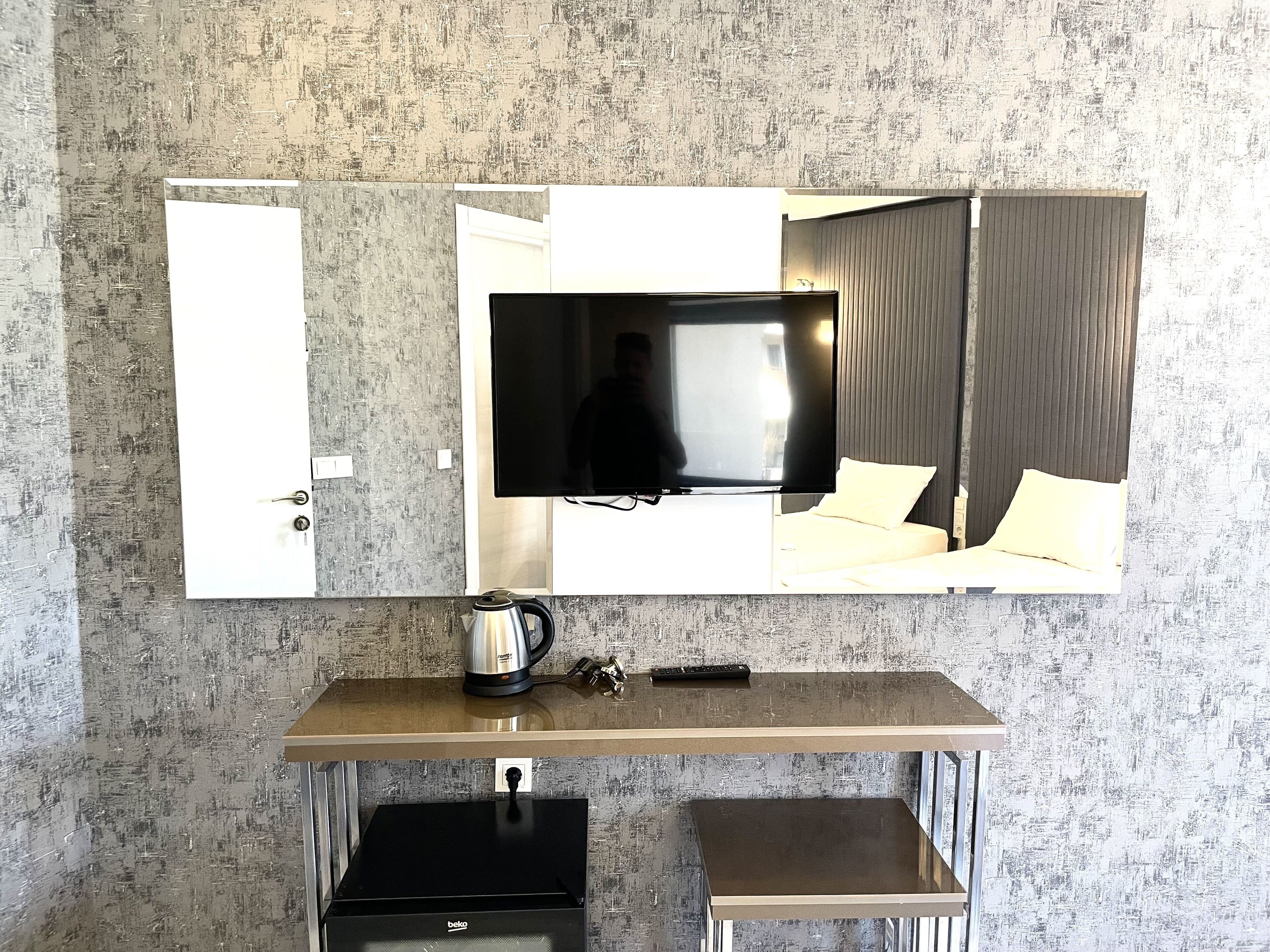 Een compacte hotelkamer met een grote spiegel, een flatscreen televisie, een kleine kitchenette met waterkoker en een bed met wit beddengoed.