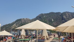 Una playa con filas de tumbonas y sombrillas, un telón de fondo montañoso y cielo despejado.