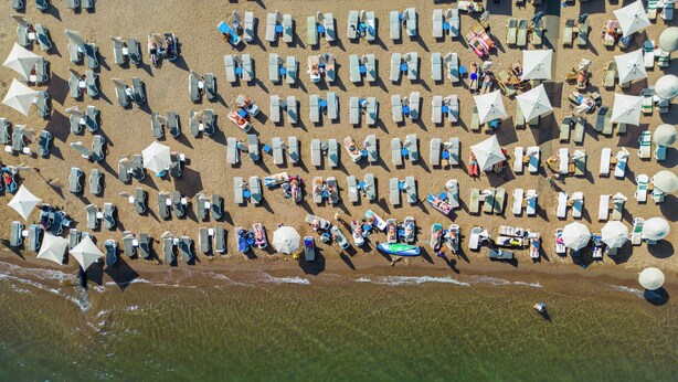 Vista aérea de una playa con filas de tumbonas y sombrillas, con una vista despejada del océano y algunas personas en el agua.