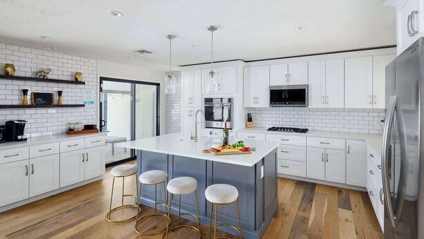 Cozinha moderna com armários brancos, uma ilha central com banquetas, eletrodomésticos de aço inoxidável e piso de madeira.
