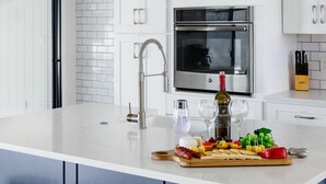 Uma cozinha moderna com armários brancos, um forno de aço inoxidável, uma garrafa de vinho e um prato de frutas na ilha.