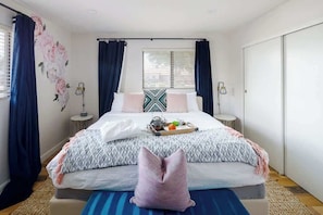 Un dormitorio con cama, cortinas, una mesita de noche y una ventana con persianas, además de un diseño floral en la pared.