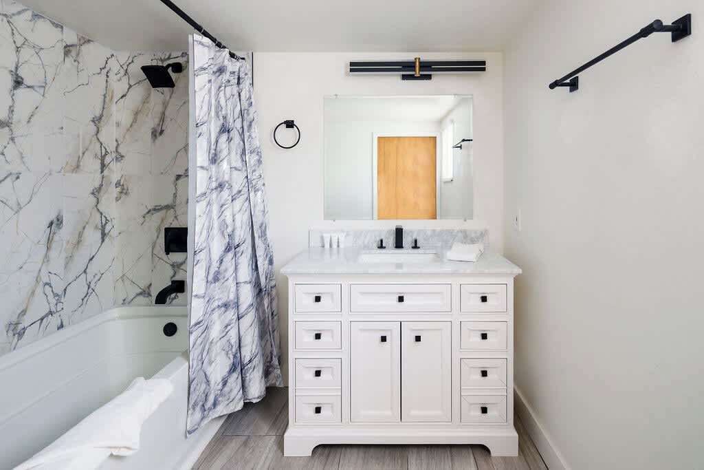 A bathroom with a marble wall, a white vanity with multiple drawers, a mirror, and a shower curtain.