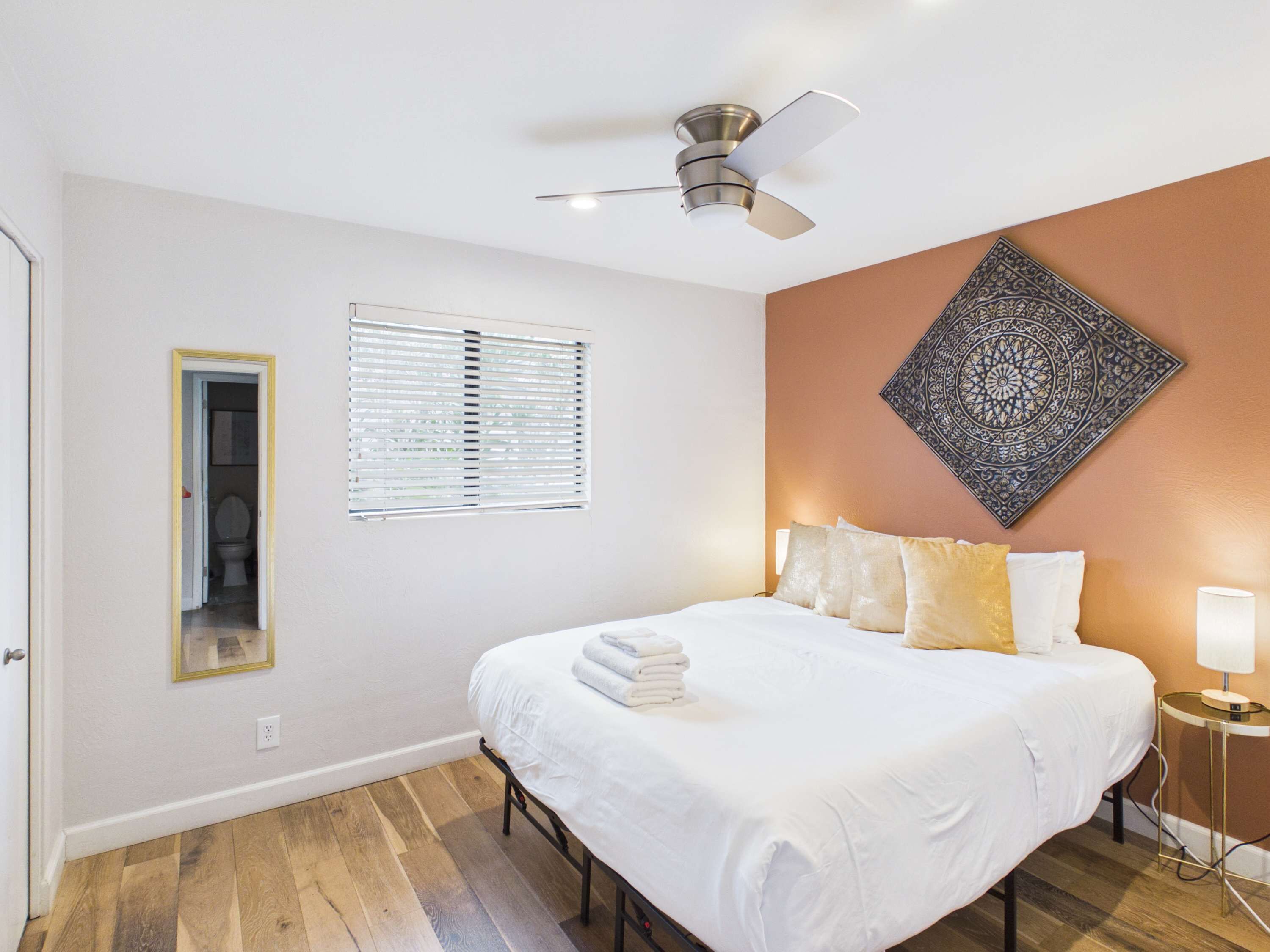 Une chambre à coucher comprenant un lit, un ventilateur de plafond, une fenêtre avec des stores, une tapisserie murale et une lampe de chevet.
