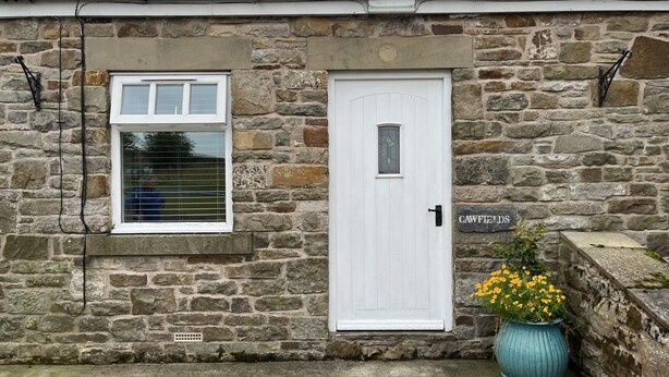 A stone building with a white door and window, a sign reading "CAWFIELD," and a potted plant with yellow flowers.
