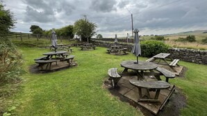 An outdoor seating area with picnic tables and umbrellas, surrounded by stone walls and a grassy field.