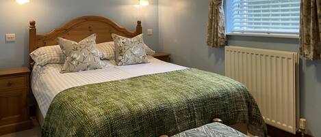 Una camera da letto con un letto in legno, cuscini floreali, una coperta verde con trama, un comodino, una lampada a parete, una finestra con tende e un termosifone.